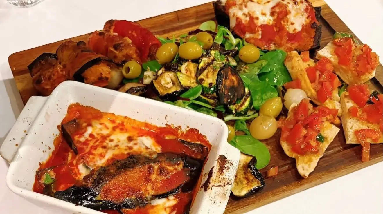 Board of Sicilian vegetable antipasti with olives, aubergine and bruschetta, beside a dish of Parmigiana