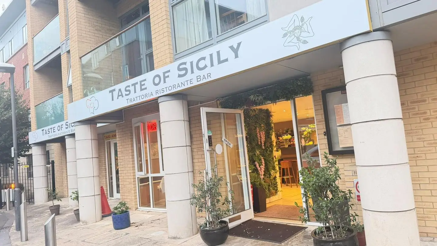 The Taste of Sicily shopfront under a yellow-brick building, the door open onto a warm-lit entrance